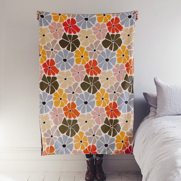 The image shows a full length blanket being held up. The design on the blanket has beautiful flowers hand sketched in autumnal tones of red, yellow, pink, green and blue.