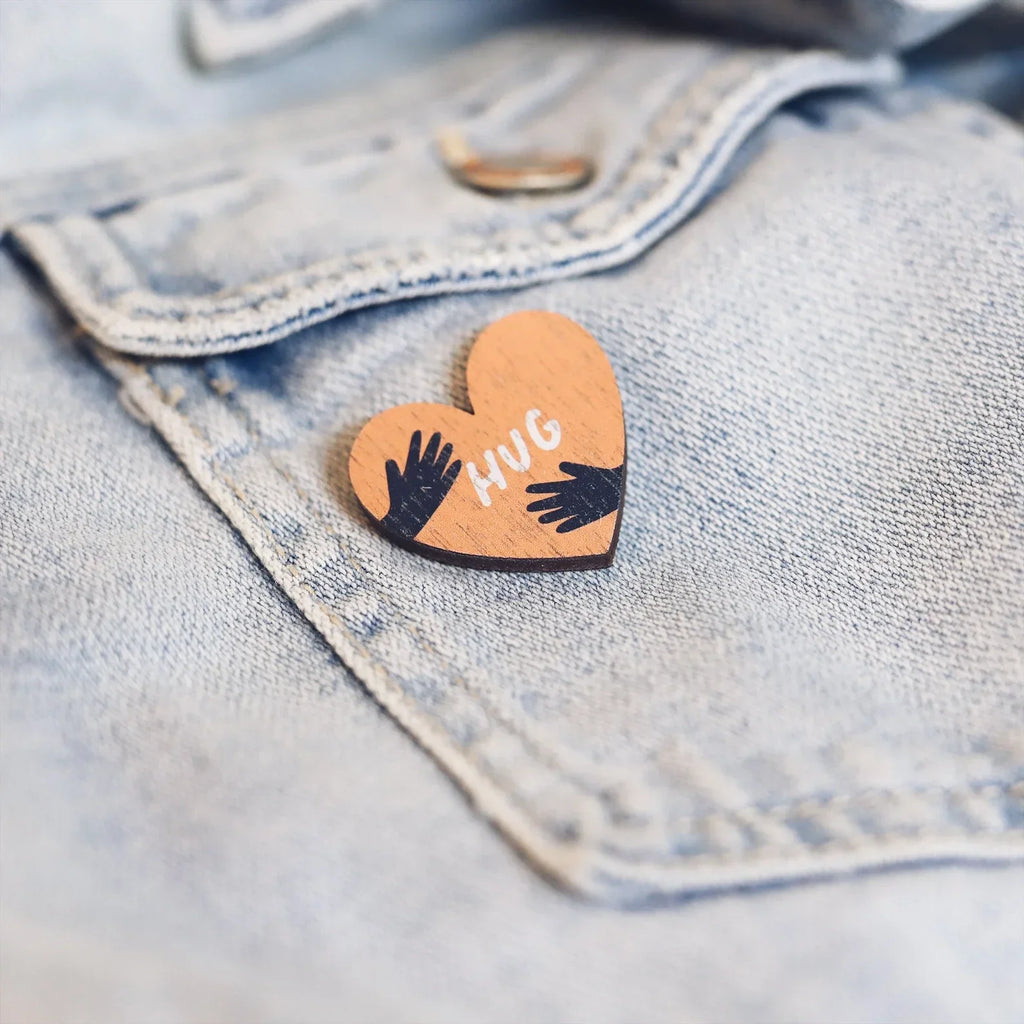 Wooden pin badge in the shape of a heart with hands wrapping round either side and the word hug in the centre. Printed on walnut veneer, pinned onto a a blue denim jacket pocket.