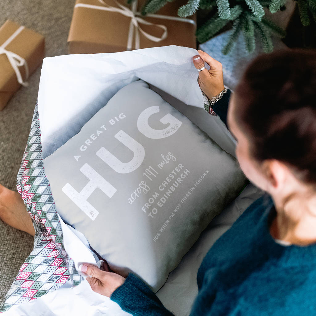 The image shows a woman unwrapping a Christmas present, inside is a personalised cushion stating the words ' a great big hug across 191 miles from Chester to Edinburgh, for when I'm not there in person x'.