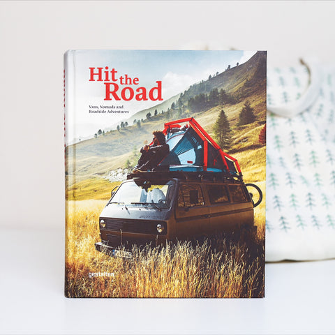A large hardback book called "Hit the Road" featuring a Volkswagen campervan with a rooftop tent. A man is sitting on the roof of the van, looking out at the tree studded hills surrounding him. 