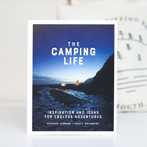 A book named "The Camping Life" with a sub title that reads "Inspiration and ideas for endless adventures". Image on the front cover is of a blue night sky, a silhouette of a mountain and a tent lit from the inside. 