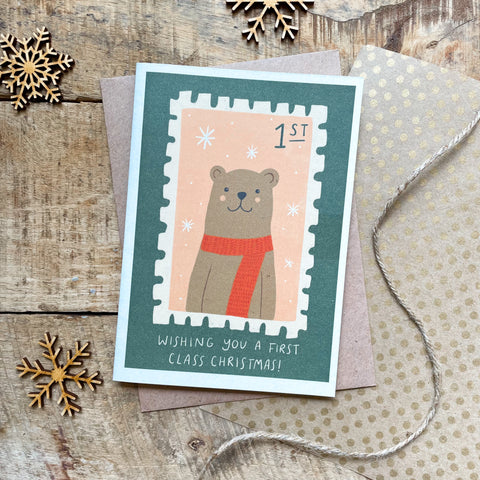 A festive card with the image of a bear wearing a red scarf on a postage stamp on a forest green background. The words "WISHING YOU A FIRST CLASS CHRISTMAS!" appear at the bottom of the card.