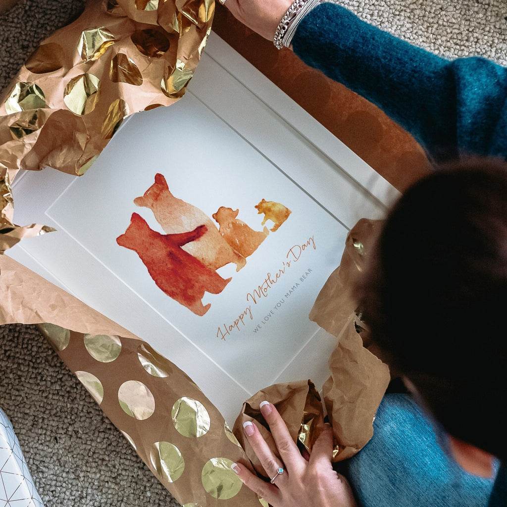 A personalised Christmas gift is being unwrapped. Inside is a custom family bear print in a black frame: The watercolour style illustrated family of bears show two parent bears, a child, and a baby bear.