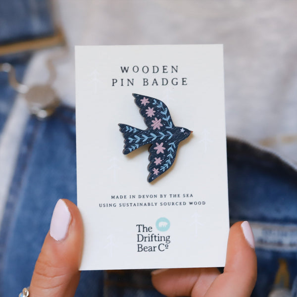 Wooden pin badge in the shape of a dove with pretty floral and botanical detail in shades of blue and pink. Printed on walnut veneer, pinned onto a recycled backing card which reads 'Wooden Pin Badge' in large text and 'Made in Devon by the sea using sustainably sourced wood' in smaller text below.