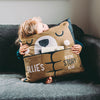A child is sat on a sofa with his arms wrapped around a cushion. The personalised Childs cushion is designed to look like a bear with the words 'Ollie's Bedtime Story Bear' written on the front. There is a pocket on the front of the cushion and we can see the top of a book poking out.
