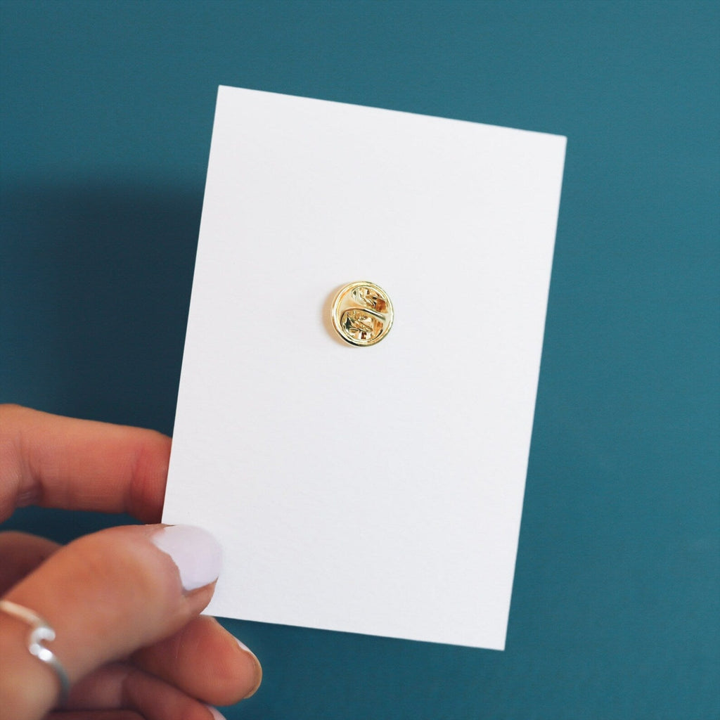 The back of a pin badge. A gold clasp attached to a recycled backing card.