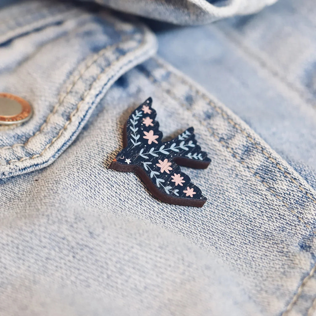 Wooden pin badge in the shape of a dove with pretty floral and botanical detail in shades of blue and pink. Printed on walnut veneer, pinned onto a blue denim jacket pocket.