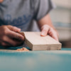 The image shows a close up of a man sanding down a solid oak wood block.