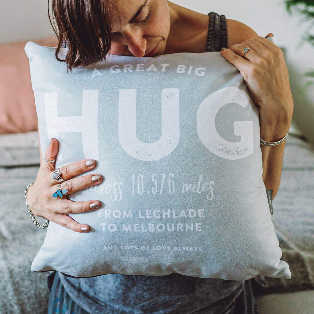 The image shows a woman holding on tight to a pebble grey coloured personalised cushion. The cushion has the words 'A Great Big HUG across 10,576 miles from Lechlade to Melbourne, and lots of love always written in bold typographic font across the front.