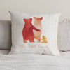 A cushion sits upon a bed with pale grey sheets. On the cushion is a family of orange bears, with two parents and a baby. Beneath the bears are the words 'The Smith Family'